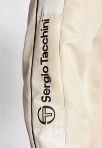 Chaqueta de color crema con una superficie texturizada, con la marca negra "Sergio Tacchini" a lo largo de la manga y un logotipo circular con iniciales.