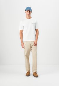 Young man wearing a white Belstaff t-shirt, beige pants, tan shoes, and a blue cap standing against a plain white background.