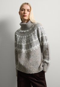 Pull à col roulé en tricot gris clair avec une bande à motif blanc, tissu texturé avec des éclats colorés, et des poignets et un ourlet côtelés.