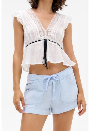 Femme portant un haut court sans manches blanc orné de dentelle, attaché par un lacet noir, et un short bleu clair à cordon de serrage avec poches.
