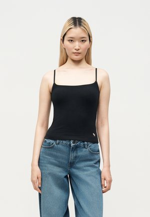 Giovane donna con capelli biondi lisci che indossa un top nero con spalline sottili e jeans blu larghi, in piedi davanti a uno sfondo semplice.