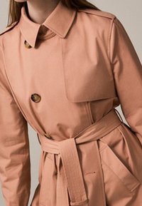 Vaalean persikan värinen trench-coat, joka on valmistettu sileästä kankaasta. Siinä on kaulus, kaksirivinen etuosa, vyö vyötäröllä ja läppätaskut.