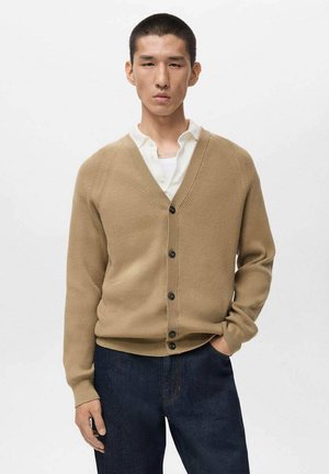 Beige ribbed cardigan with a V-neck, six dark buttons down the front, and long sleeves, worn over a white shirt and dark blue jeans.