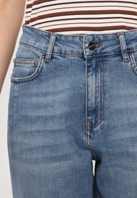 Jeans de mezclilla de color azul claro con una cintura alta, cinco bolsillos y costuras naranjas contrastantes. Presenta un cierre con botón metálico y cremallera.