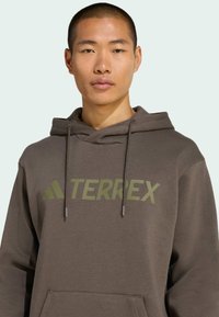 Sudadera marrón con un gran logo "TERREX" en verde oliva, cordones y un bolsillo canguro, hecha de una suave y texturizada tela.