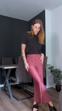 Black t-shirt, pleated pink trousers, and black sandals. Natural wood floor, dark wall background, and green plant in a pot.