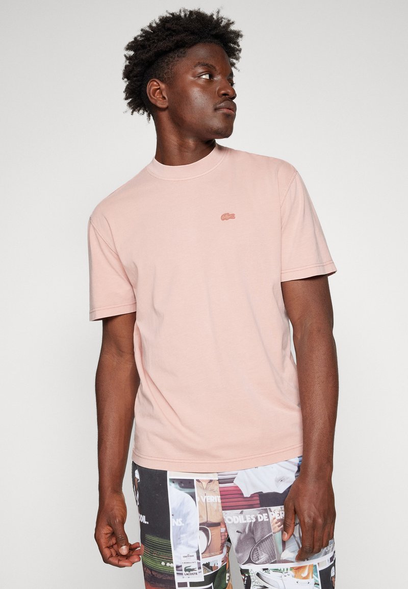 T-shirt en coton rose clair avec un col rond et des manches courtes. Présente un petit logo discret au centre de la poitrine. Texture lisse.