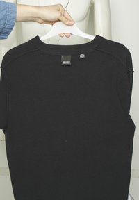 Pull noir à côtes avec un col rond et des manches courtes, comportant une étiquette au dos. Le tissu a une finition texturée.