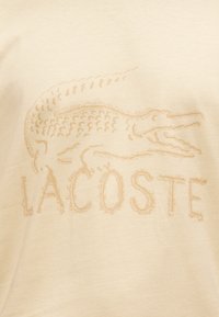 Μπεζ βαμβακερό μπλουζάκι με υφασμένο λογότυπο κροκοδείλου και τη λέξη "LACOSTE" σε ελαφρώς πιο σκούρο τόνο, τοποθετημένα στο κέντρο του μπροστινού μέρους.