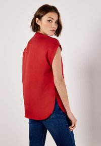 Rote ärmellose Bluse mit hohem Kragen, seitlichen Schlitzen und lockerer Passform. Glatte Textur, fließender Stoff und leicht abgerundeter Saum.