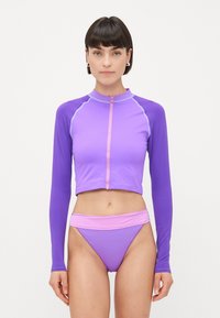 Sieviete, ģērbusies purpura ilga piedurkne ar rāvējslēdz�ēju un atbilstošiem augstiem bikini apakšbiksēm ar gaiši rozā jostasvietu, stāv uz balta fona.