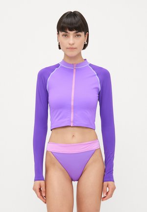 Mujer vestida con un top corto de manga larga y cremallera color púrpura y una bikini de corte alto a juego con una cinturilla rosa claro, de pie contra un fondo blanco.