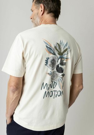 Uomo con barba grigia che indossa una maglietta bianco sporco con un design floreale in stile pittura a mano e la scritta "Mind in Motion" sul retro.
