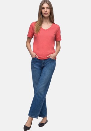 Frau trägt korallenfarbene Kurzarm-V-Ausschnitt-Hemd, blaue Jeans und schwarze Ballerinas, steht mit den Händen in den Taschen vor schlichtem Hintergrund.