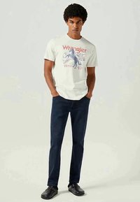 Hvit t-skjorte med rød og blå Wrangler-grafikk, kombinert med mørkeblå denim-bukser og svarte sko, stående på en nøytral bakgrunn.
