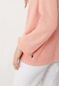 Jasnoróżowy sweter dzianinowy z teksturowanym wzorem, z okrągłym dekoltem i podwiniętymi rękawami. Mała czarna metka z logo w pobliżu brzegu.