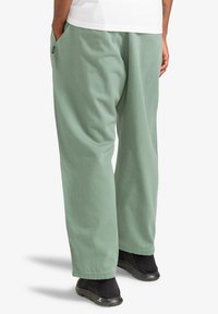 Man in ruime groene broek en zwarte instappers, staand met handen in zijn zakken, gezien vanuit de achterkant tegen een witte achtergrond.