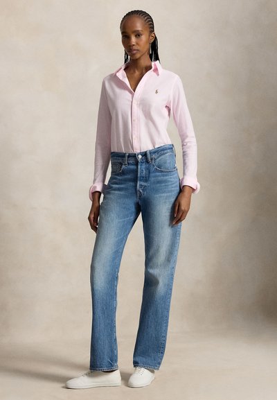 Giovane con capelli intrecciati che indossa una camicia rosa chiaro con bottoni, jeans blu e sneakers bianche, in piedi su uno sfondo semplice.