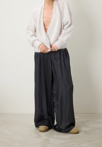 Vaaleanharmaa pörröinen kardigan persikanvärisen toppikujan päällä, yhdistetty mustiin satiinisiin leveisiin housuihin ja beigeisiin pörröisiin tossuihin beigen lattian päällä.