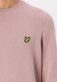 Lichtroze gebreide trui met een ronde hals en geribbelde manchetten, met een klein zwart en gouden vogel logo op de borst.