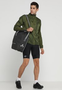 Giacca leggera verde con zip frontale, borsa nera impermeabile con cinghie grigie e pantaloni corti da ciclismo neri aderenti.