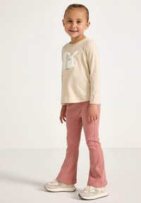 Beige langærmet skjorte med tekstureret hvid kanindesign, parret med pink ribbede bukser. Modellen bærer hvide og grå sneakers.