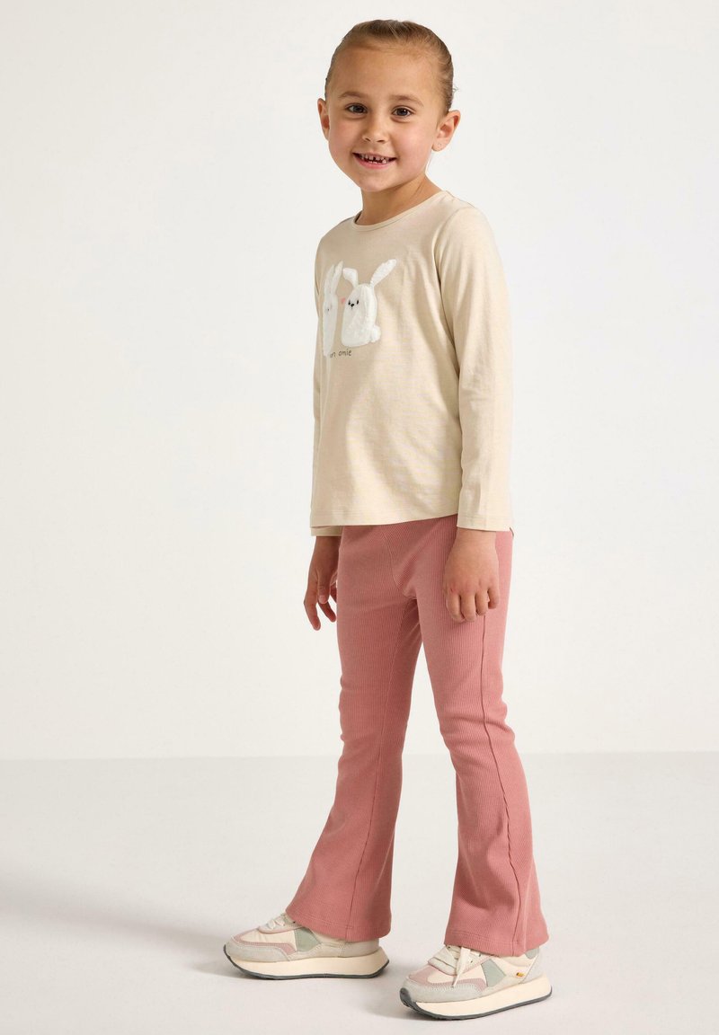 Beige langærmet skjorte med tekstureret hvid kanindesign, parret med pink ribbede bukser. Modellen bærer hvide og grå sneakers.