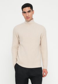 Mann in beigem geripptem Rollkragenpullover und schwarzen Hosen, der mit einer Hand in der Tasche vor einem weißen Hintergrund steht.