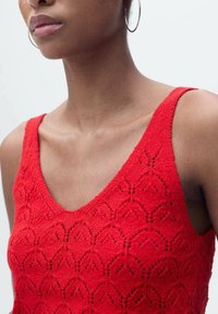 Top in maglia rossa a canotta con scollo a V, caratterizzato da un motivo testurizzato a forma di diamante e dettagli a giorno lungo il tessuto.
