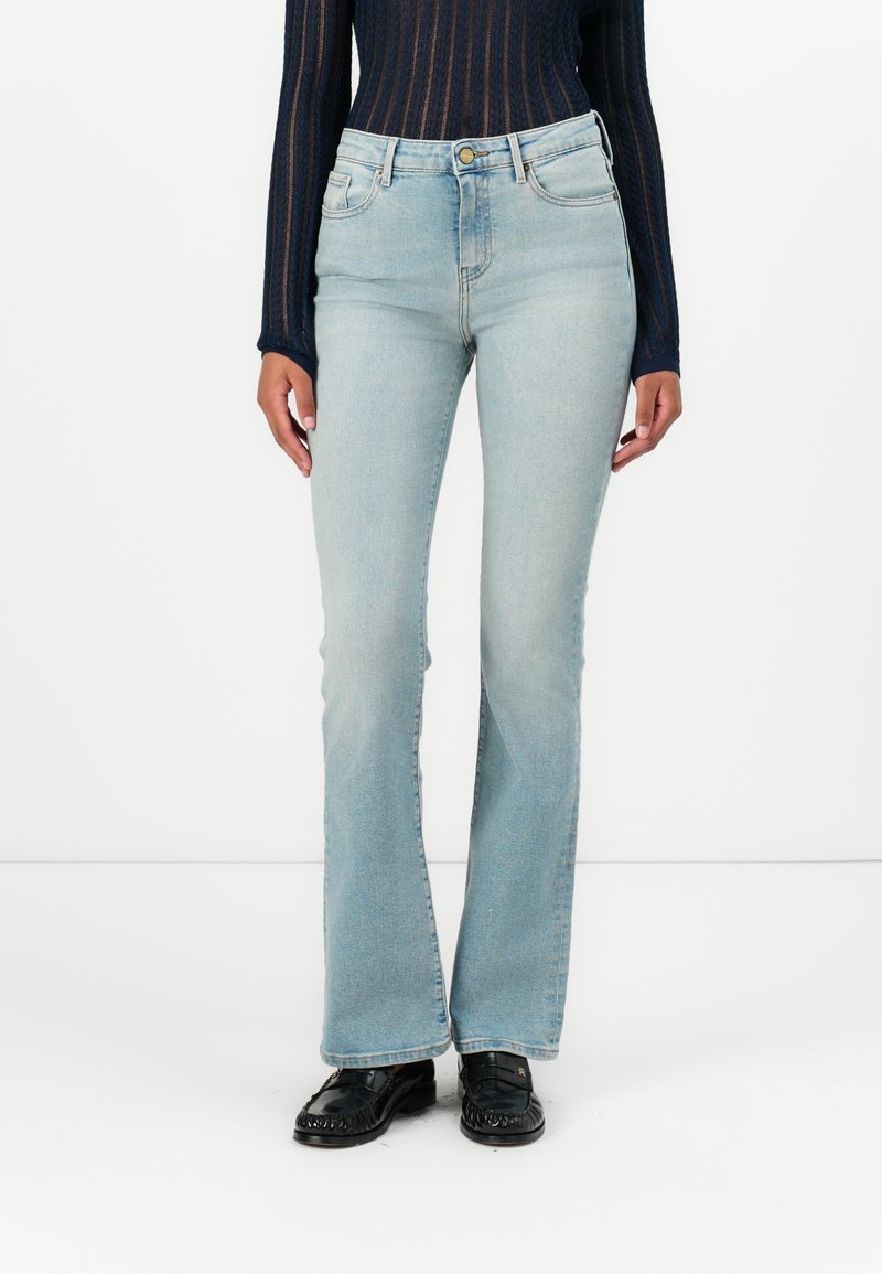 Γυναίκα με ανοιχτό μπλε τζιν παντελόνι bootcut, μαύρα μοκασίνια και μακρυμάνικη σκουρόχρωμη ριγωτή μπλούζα, που στέκεται μπροστά σε λευκό φόντο.