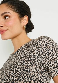 Haut à imprimé léopard avec des manches courtes, présentant un fond beige et des taches noires. Boucle d'oreille en forme de créole texturée dorée qui complète la tenue.