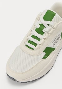 Krémszínű hálós felsőrészű sneaker, zöld kiemelésekkel és fehér fűzőkkel. Texturált velúr orrész és gumiós aljzattal rendelkezik. Márkajelzés az oldalon.