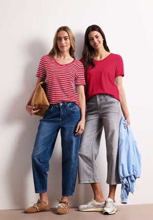 Two women standing against a white wall, one in red striped t-shirt and jeans holding a brown bag, the other in plain red t-shirt and gray pants holding a blue jacket.