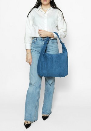 Sac bandoulière en daim bleu avec corps arrondi, anse supérieure et détails cousus ; porté par une personne vêtue d'une chemise blanche et d'un jean en denim.