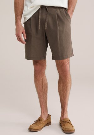 Barna zsinóros rövidnadrágot, fehér inget és bézs színű velúr mokaszint viselő férfi áll egyszínű háttér előtt.