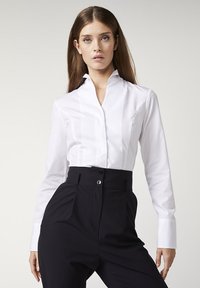 Weiße langärmlige Bluse mit hohem Kragen, vertikalen Details und glatter Textur, kombiniert mit hoch taillierten schwarzen Hosen.