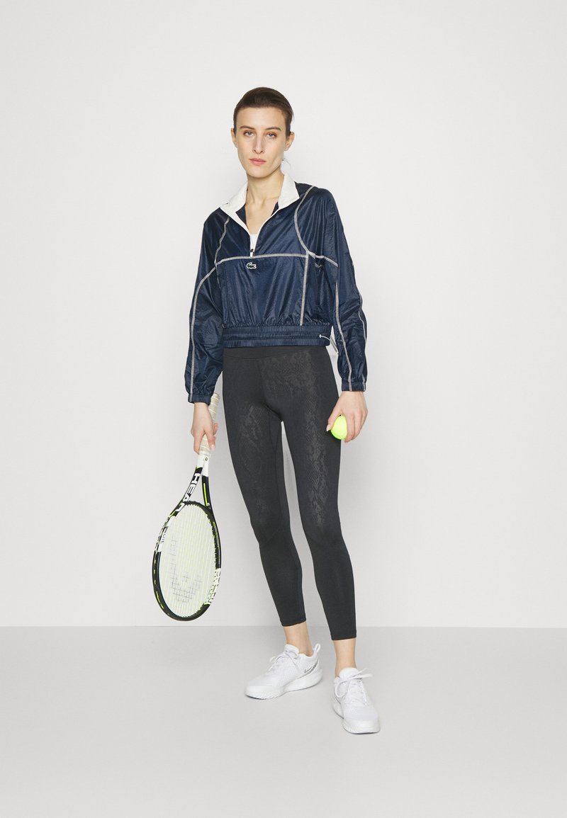Veste cropped bleue marine avec col blanc et coutures grises, portée avec des leggings noirs à motif subtil, tenant une raquette de tennis et une balle.