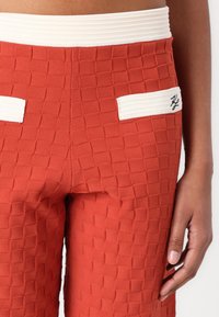 Gelaagde, baksteenkleurige, roestkleurige broeken met een witte elastische tailleband en zakken, voorzien van een klein geborduurd logo op de rechterzak.