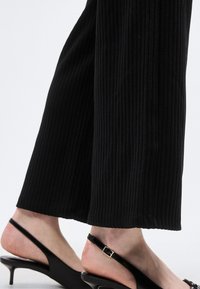 Κοντινό πλάνο των κάτω άκρων με μαύρο παντελόνι με ριμπ ύφανση και μαύρα γοβάκια με λουράκι στον αστράγαλο, λεπτά λουράκια και μικρές αγκράφες.