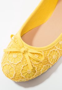 Ballerine jaune avec motif en dentelle florale et petit nœud noué sur l'orteil, montrant une doublure intérieure beige douce.