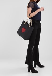 Svart tote-väska med ett röd hjärtformad design, guld kedjehandtag och en slät textur. Väskan har en strukturerad form och är rymlig.
