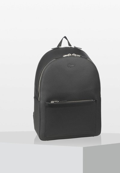 Sac à dos noir en matériau texturé, avec un dessus arrondi, une poche avant zippée et des accents en métal argenté. Logo Lacoste embossé à l'avant.