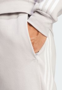 Lys grå, bløde joggers i bomuldsblanding med sidelomme. Har hvide stribedetaljer langs siden. Tekstureret stof synligt.