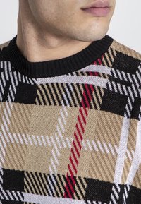 Mønstret sweater med et ternet design i beige, sort, hvid og rød med en ribbet sort halsudskæring og struktureret strikket stof.