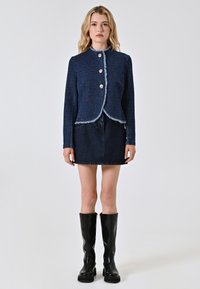 La modella si erge con sicurezza indossando una giacca cropped blu scuro con bottoni argentati, abbinata a una gonna di jeans e stivali neri alti, su uno sfondo semplice.