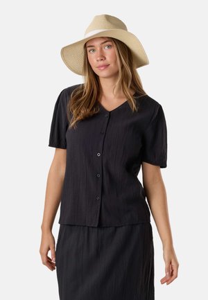Femme aux cheveux châtain clair portant un chapeau beige à large bord et un chemisier noir texturé à manches courtes boutonné et une jupe.