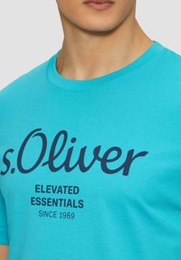 Turquoise cotton t-shirt with round neckline. Features black printed text: "s.Oliver ELEVATED ESSENTIALS SINCE 1969" prominently displayed.