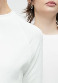 Valge soonikkoega topp ümmarguse kaeluse ja raglaanvarrukatega, esitatud hele tausta ees, iseloomustatuna siledast tekstuurist ja peentest detailidest.