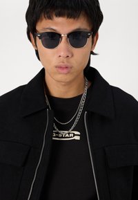 Μαύρο μπουφάν από μείγμα μάλλινου υφάσματος με φερμουάρ, επενδεδυμένο πάνω από μια μαύρη μπλούζα με λευκό λογότυπο, συμπληρωμένο με ασημένια αλυσίδες και σκούρα γυαλιά ηλίου.