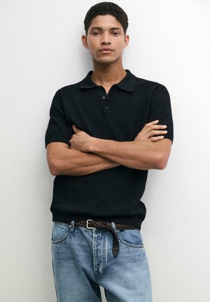 Junger Mann mit kurzen lockigen Haaren steht mit verschränkten Armen, trägt ein schwarzes kurzärmeliges, geknöpftes Hemd und hellblaue Jeans mit geflochtenem Gürtel.